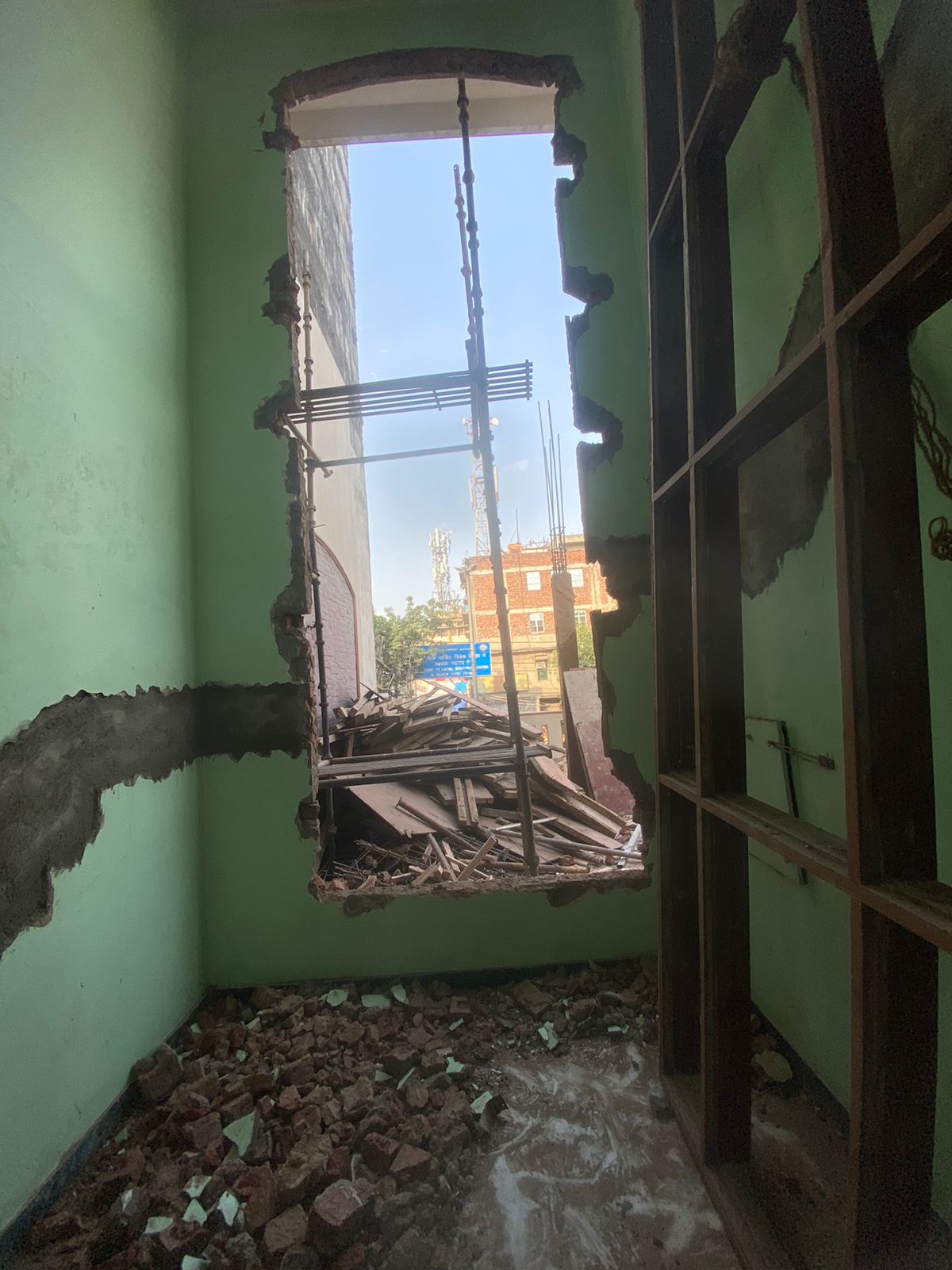 A construction room showing a large cut-out window opening. On the right, a large unused teakwood frame leans against the wall amidst construction debris.